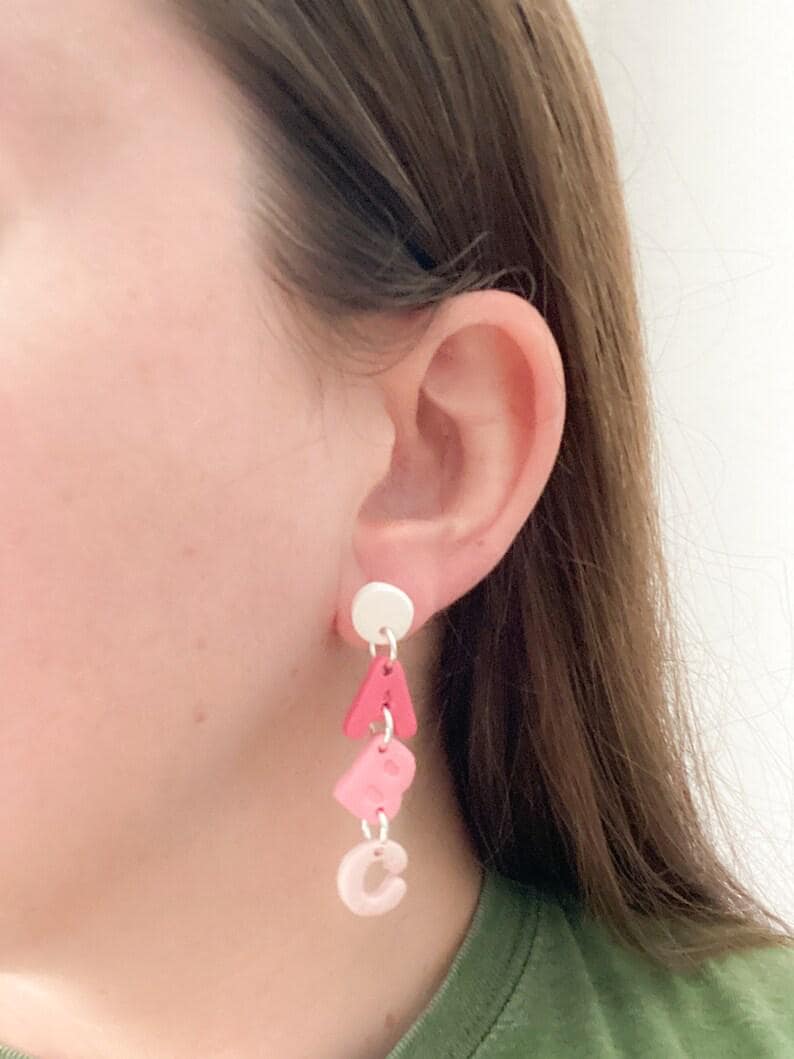 May include: A dangle earring with a white stud and three pink letter charms spelling "ABC". The letters are in varying shades of pink, with the letter "A" being a darker shade than the other letters.