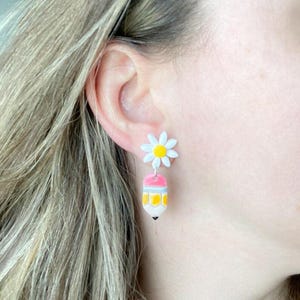 May include: Daisy and pencil-shaped earrings. The earrings feature a white daisy with a yellow center and a pink, yellow, and white pencil design. The pencil has a black tip. The earrings are hanging from a person's ear.