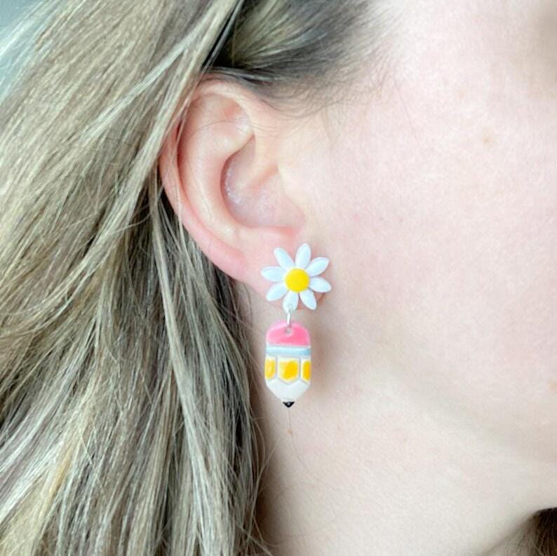 May include: Daisy and pencil-shaped earrings. The earrings feature a white daisy with a yellow center and a pink, yellow, and white pencil design. The pencil has a black tip. The earrings are hanging from a person's ear.