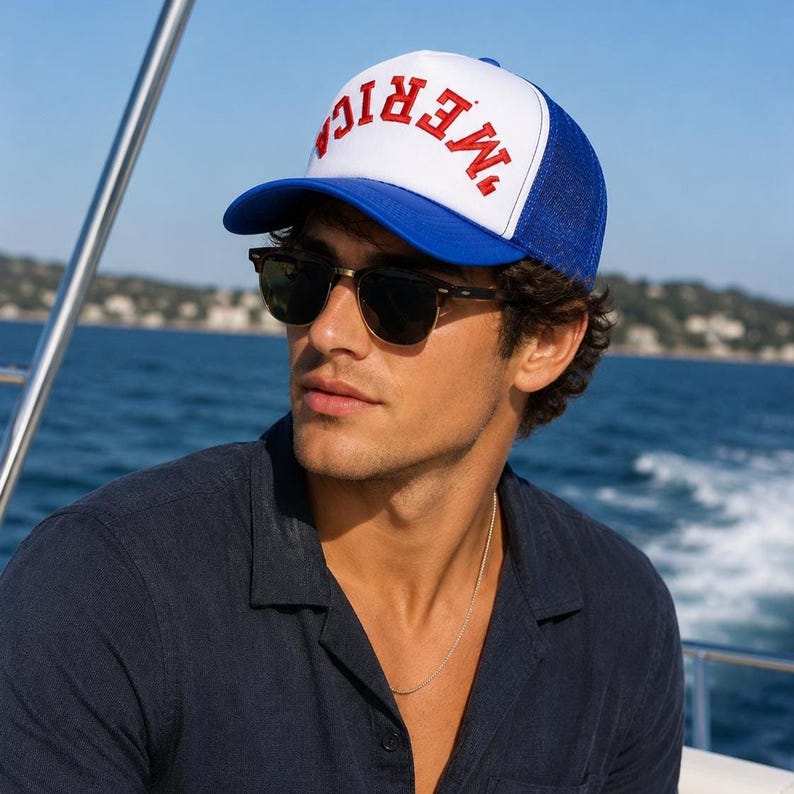 May include: A white and blue trucker cap featuring the red text MERICA printed in a bold, arched font across the front panel. The cap has a solid white front, a blue brim, and a blue mesh back panel with an adjustable snap closure at the rear.