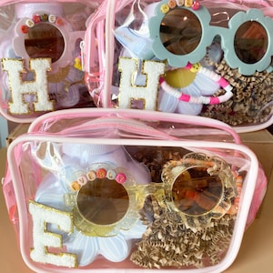 May include: Clear pink zippered cosmetic bags filled with various items. Each bag features a large initial, a pair of flower-shaped sunglasses, and a bottle with a name on it. The bags contain bracelets and other accessories.