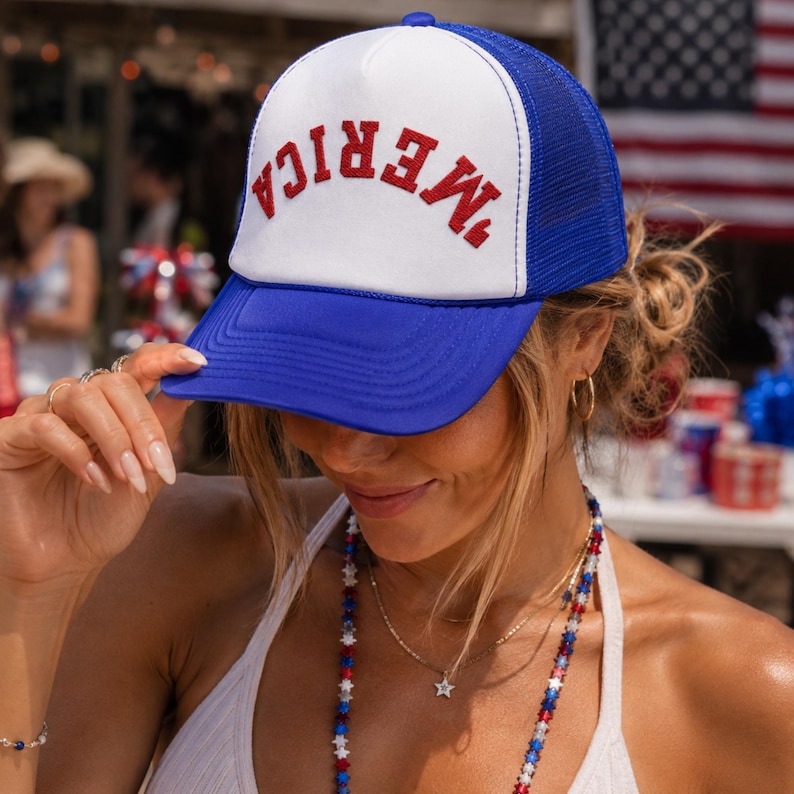 May include: A blue and white trucker cap featuring a mesh back and a solid front panel. The word 'MERICA' is embroidered in red, arched lettering across the centre of the white front panel. The cap has a solid blue brim and a blue mesh back.