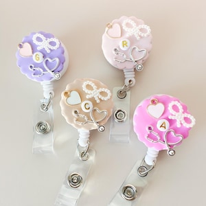 May include: Three colorful retractable badge holders with a heart, stethoscope, and bow design. The holders are pink, purple, and beige with gold accents. The letters "G", "C", and "A" are visible on the holders.