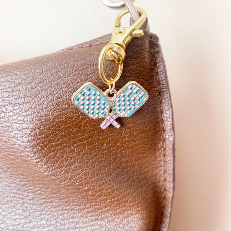 May include: A gold-colored keychain clip holding a charm with two crossed pickleball paddles. The paddles are light blue with white grid patterns and a lavender handle. The charm hangs against a brown leather bag.