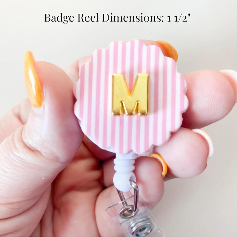 Puede incluir: Un portacredenciales redondo con bordes festoneados, un patr&oacute;n de rayas verticales rosas y blancas y una letra M dorada centrada en la parte frontal. El texto Badge Reel Dimensions: 1 1/2" es visible en la parte superior.
