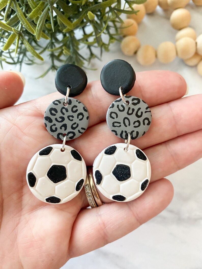Może przedstawiać: Para kolczyk&oacute;w wiszących w kolorze czarnym, szarym i białym. Kolczyki mają wisiorki w kształcie piłki nożnej, szary wisiorki z nadrukiem lamparta i czarny wisiorki.