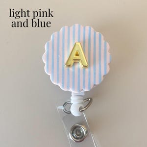 Puede incluir: Un portatarjetas con una cara circular festoneada con rayas verticales de color rosa claro y azul, con una letra A dorada centrada en el frente. El texto light pink and blue est&aacute; impreso en la esquina superior izquierda de la imagen.