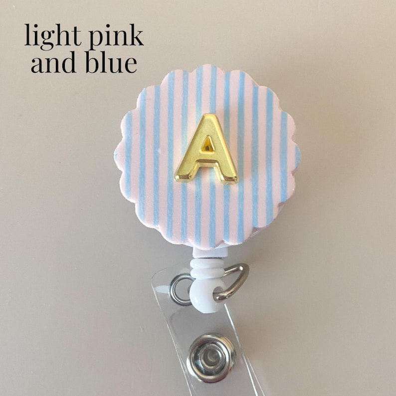 Puede incluir: Un portatarjetas con una cara circular festoneada con rayas verticales de color rosa claro y azul, con una letra A dorada centrada en el frente. El texto light pink and blue est&aacute; impreso en la esquina superior izquierda de la imagen.