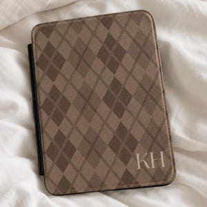 May include: A rectangular tablet or e-reader case featuring a brown and tan argyle diamond pattern. The front cover displays the white capital letters KH in the bottom right corner. The case has a solid black spine and a textured exterior finish.