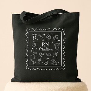 May include: Black tote bag featuring a white square design with medical-themed illustrations and the text "RN Madison." The design includes a stethoscope, heart, and medical tools. The bag has two handles for carrying.