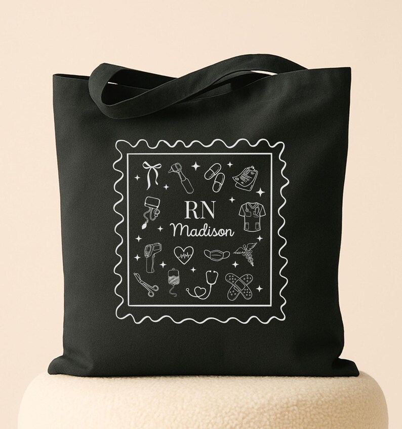 May include: Black tote bag featuring a white square design with medical-themed illustrations and the text "RN Madison." The design includes a stethoscope, heart, and medical tools. The bag has two handles for carrying.