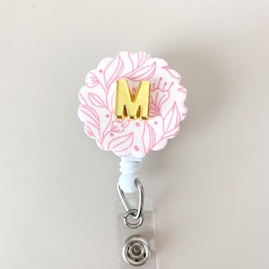 May include: A floral-patterned badge reel with a gold letter "M" in the center. The badge reel has a white base with pink floral designs. The reel is attached to a white retractable cord and a clear plastic clip.