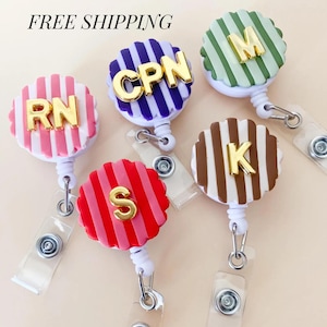 May include: Five circular badge reels with scalloped edges, each featuring vertical stripes in pink, purple, green, red, or brown. Each reel displays gold-colored letters including RN, CPN, M, S, and K. The reels have white plastic housings and clear vinyl straps with metal snaps.