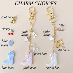 May include: A gold chain with charms, including a pink heart, a white heart, a gold bow, a pearl bow, a letter F, a blue heart, a pink boot, a cream boot, a gold boot, a cherry, and the letter A. The charms are labeled with their names.
