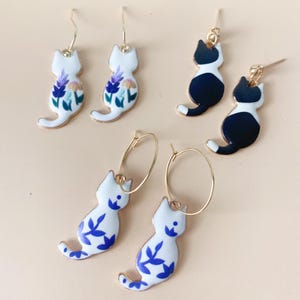 May include: Three pairs of cat-shaped earrings. One pair is white with purple flowers, one pair is black and white, and one pair is white with blue floral designs.