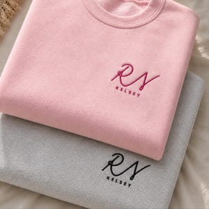 Pode incluir: Uma camisola rosa claro e uma camisola cinzenta empilhadas uma sobre a outra. Cada camisola apresenta um logótipo bordado com as letras estilizadas RS e o nome KELSEY por baixo. A camisola rosa tem um bordado em rosa escuro.