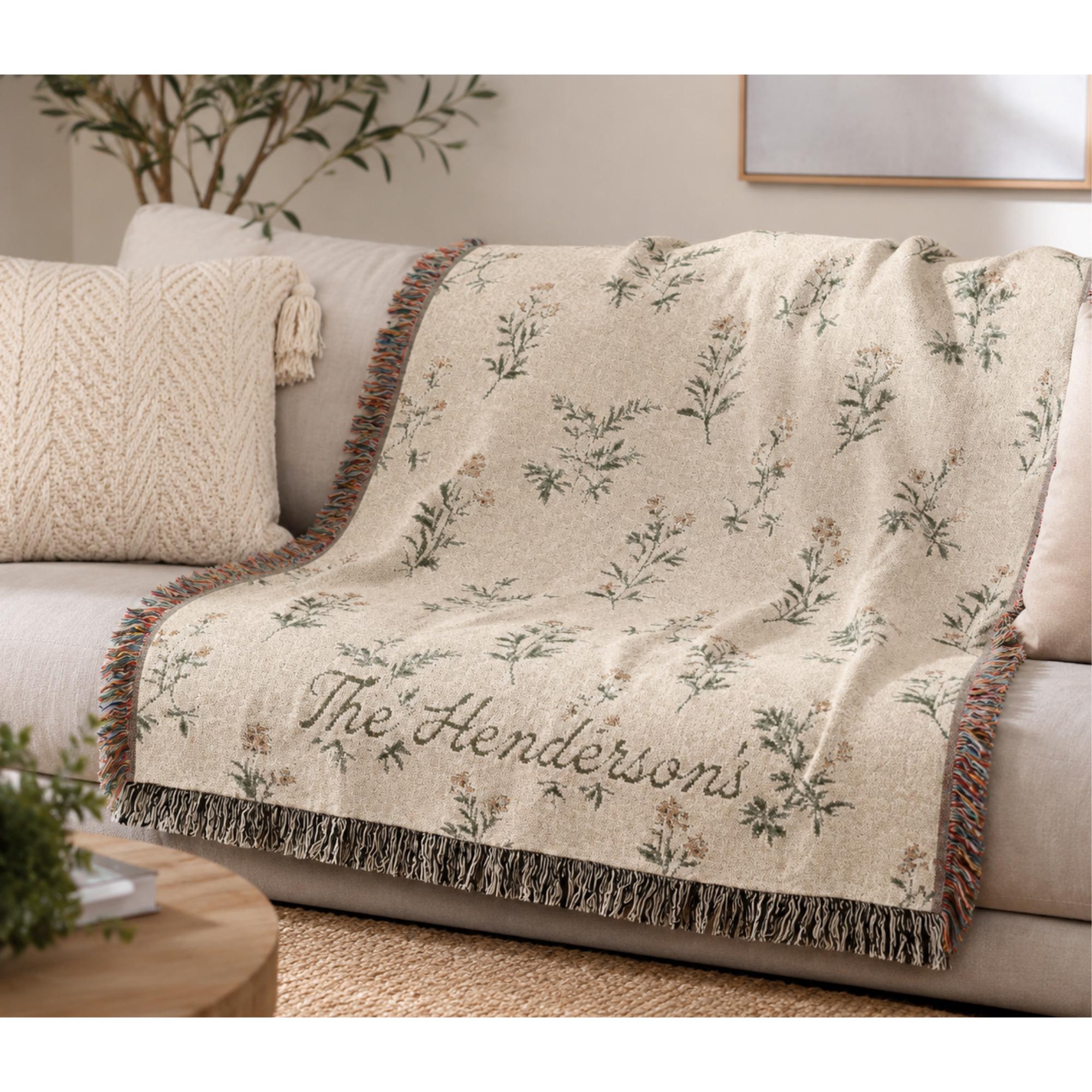Pode incluir: Uma manta tecida de cor creme com um padr&atilde;o repetitivo de ilustra&ccedil;&otilde;es bot&acirc;nicas florais em verde e castanho. O texto "The Henderson's" est&aacute; bordado em letra cursiva. A manta tem extremidades com franjas de fios multicoloridos.