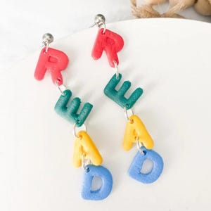 May include: A pair of colorful dangle earrings spelling out the word "READ DAD". The earrings feature red, green, yellow, and blue letters, suspended from silver-colored posts. The letters are in a playful, blocky font.