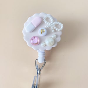 May include: A white and pink badge reel with a pill, smiley face, and pearl accents. The reel has a silver clip and a retractable cord. The badge reel is decorated with a pearl bow and a gold letter "P".