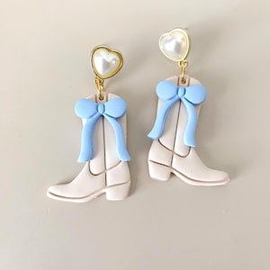 May include: A pair of earrings featuring a cowboy boot design. The earrings are off-white with a light blue bow and a gold-colored heart-shaped top with a pearl-like center. The earrings are on a light beige background.