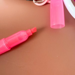 Puede incluir: Un marcador fluorescente de color rosa brillante con tapa extra&iacute;ble y punta biselada. El marcador est&aacute; unido a una anilla de pl&aacute;stico rosa y a un mecanismo de clip negro, presentando un dise&ntilde;o compacto para facilitar su transporte y uso.