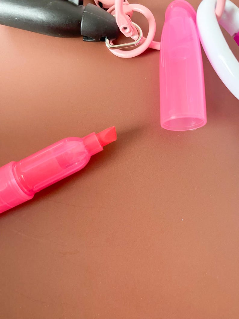 Puede incluir: Un marcador fluorescente de color rosa brillante con tapa extra&iacute;ble y punta biselada. El marcador est&aacute; unido a una anilla de pl&aacute;stico rosa y a un mecanismo de clip negro, presentando un dise&ntilde;o compacto para facilitar su transporte y uso.