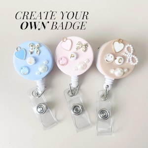 May include: Three colorful retractable badge holders with different embellishments. The badge holders are blue, pink, and beige with white accents. Each badge holder has a heart, a bow, a smiley face, and a pearl. The text "CREATE YOUR OWN BADGE" is at the top of the image.