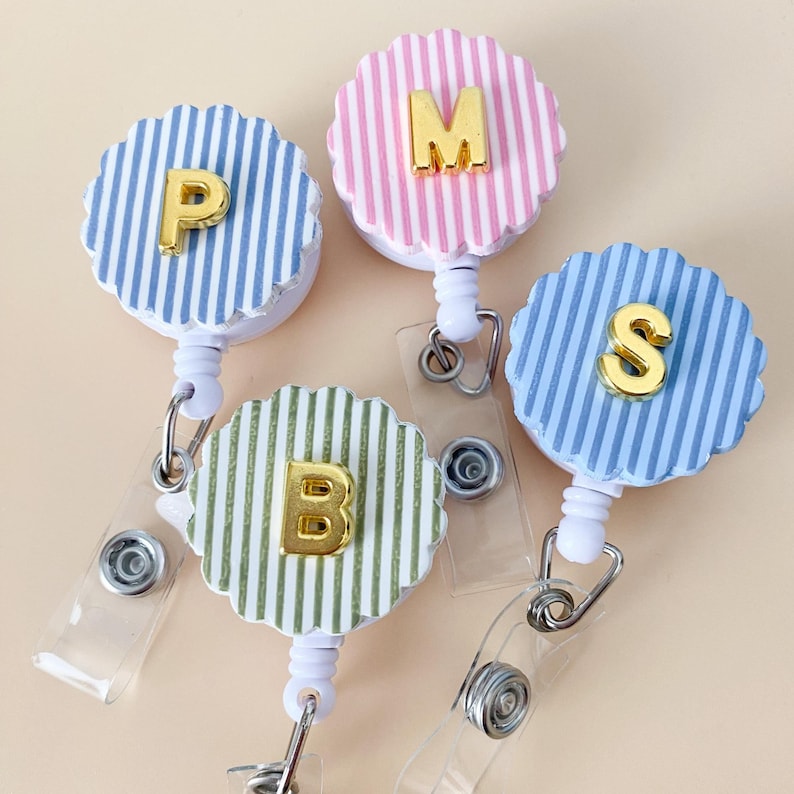 Puede incluir: Cuatro portatarjetas circulares con bordes festoneados, con rayas verticales en azul, rosa, verde y azul claro. Cada uno tiene una letra dorada met&aacute;lica en relieve: P, M, B y S. Incluyen carcasas de pl&aacute;stico blanco y correas de vinilo transparente con broches de metal.