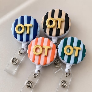 May include: Four circular badge reels with scalloped edges, each featuring vertical stripes in blue, beige, orange, or green. Each reel displays the gold-colored letters OT in the center, attached to a white plastic housing with a clear strap.