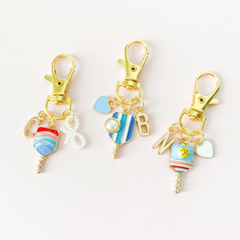 May include: Three gold keychains with blue, red, and white paddleball charms. Each charm has a small pearl, a heart, and a letter. The letters are C, B, and N.