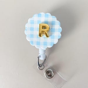 May include: A badge reel with a light blue and white gingham pattern and a gold letter "R". The reel has a scalloped edge and a clear plastic strap with a metal snap.