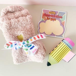 K&ouml;nnte beinhalten: Ein Paar rosa flauschige Socken, die mit einem Regenbogenband gebunden sind, ein gelber Bleistift mit einem rosa Radiergummi und eine Karte mit dem Text "With Appreciation to You" in einer Retro-Schrift.