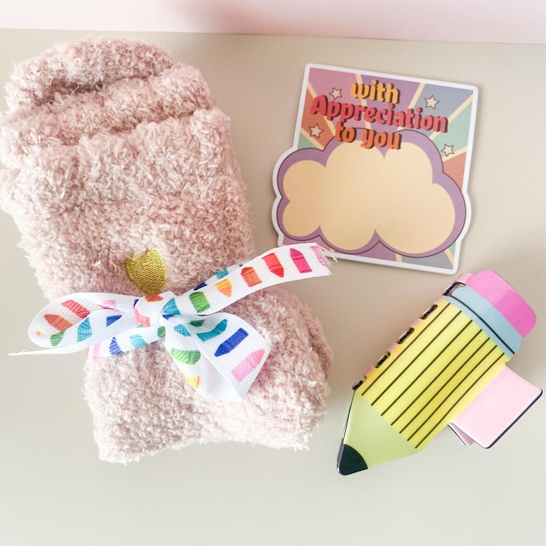K&ouml;nnte beinhalten: Ein Paar rosa flauschige Socken, die mit einem Regenbogenband gebunden sind, ein gelber Bleistift mit einem rosa Radiergummi und eine Karte mit dem Text "With Appreciation to You" in einer Retro-Schrift.