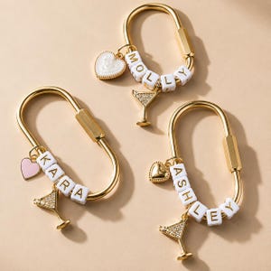 May include: Three gold-toned oval carabiner keychains featuring white cube beads spelling the names MOLLY, KARA, and ASHLEY. Each keychain includes a dangling charm: a white heart, a pink heart, or a gold heart, plus a crystal-encrusted martini glass.