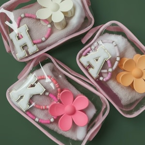 May include: Clear pink cosmetic bags containing a white towel, a beaded bracelet, a flower-shaped hair clip, and a letter charm. The letters are white with gold trim. The hair clips and bracelets are in shades of pink and white.