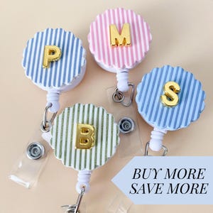 May include: Four badge reels with different colored striped backgrounds and gold letters: P, M, B, and S. The reels are white with a clear plastic strap. The text "BUY MORE SAVE MORE" is in a light blue banner.
