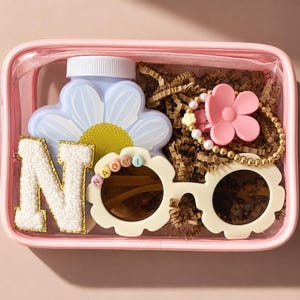 Pode incluir: Uma bolsa retangular rosa transparente contendo uma garrafa azul-clara em forma de margarida, um patch com a letra N branca e borda dourada, óculos de sol creme em forma de flor com o nome NAOMI e um prendedor de cabelo de flor rosa.