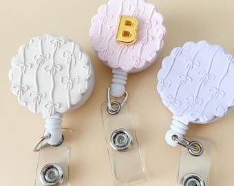 Puede incluir: Tres portatarjetas con bordes decorativos festoneados y detalles de lazo. Uno es blanco, otro es rosa claro con la letra dorada "B", y otro es lavanda. Cada carrete tiene una correa de plástico transparente y un clip de metal.