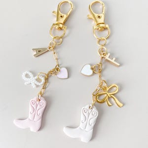 May include: Two gold keychains with charms. One keychain has a pink cowboy boot, a pink heart, a white bow, and the letter A. The other keychain has a white cowboy boot, a white heart, a gold bow, and the letter F.
