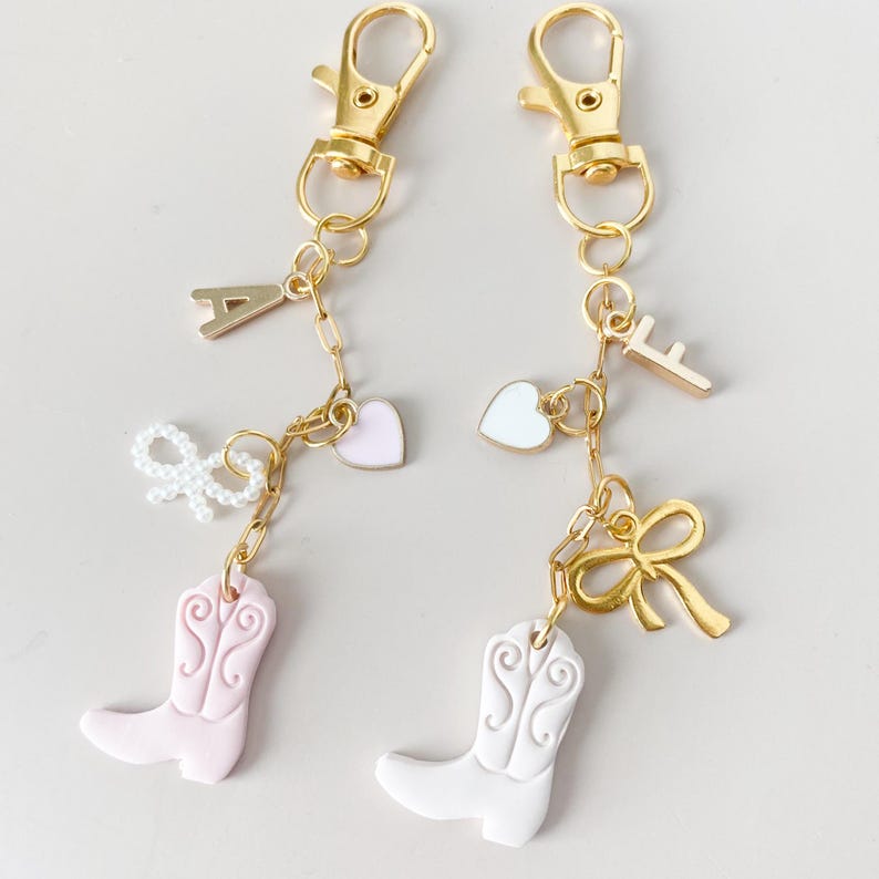 May include: Two gold keychains with charms. One keychain has a pink cowboy boot, a pink heart, a white bow, and the letter A. The other keychain has a white cowboy boot, a white heart, a gold bow, and the letter F.