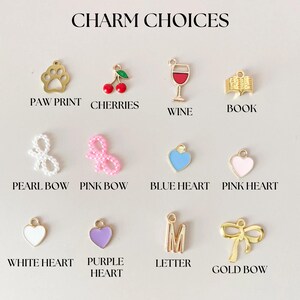 May include: A selection of charms for jewelry making, including a paw print, cherries, a wine glass, a book, a pearl bow, a pink bow, a blue heart, a pink heart, a white heart, a purple heart, a letter M, and a gold bow.