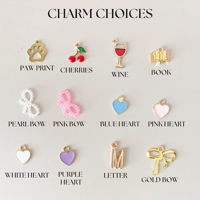 May include: A selection of charms for jewelry making, including a paw print, cherries, a wine glass, a book, a pearl bow, a pink bow, a blue heart, a pink heart, a white heart, a purple heart, a letter M, and a gold bow.