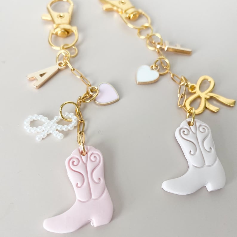 May include: Two gold keychains with charms. One keychain has a pink boot, a pink heart, a pearl bow, and the letter A. The other keychain has a white boot, a white heart, a gold bow, and the letter F.