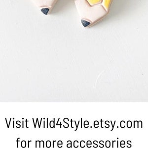 May include: A pair of whimsical pencil-shaped earrings with gold hoops. The earrings feature pink erasers, yellow pencil bodies, and black tips. The image also shows heart-shaped earrings in red, blue, and white.