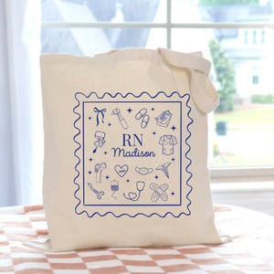 May include: A cream-colored tote bag with a blue square design featuring medical icons and the text "RN Madison." The bag has a rectangular shape with a handle for carrying. The design includes a stethoscope, heart, and other medical symbols.