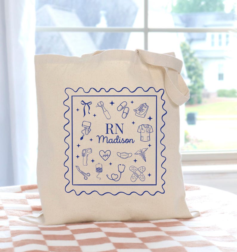 May include: A cream-colored tote bag with a blue square design featuring medical icons and the text "RN Madison." The bag has a rectangular shape with a handle for carrying. The design includes a stethoscope, heart, and other medical symbols.