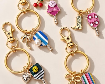 Puede incluir: Una colección de llaveros de metal dorado con varios dijes de esmalte. Los dijes incluyen paletas de pickleball, una bota vaquera, una palmera, un corazón, un lazo, cerezas y flores. Algunas paletas incluyen el texto PICKLEBALL.