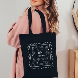 May include: Black tote bag with white line art of medical tools and the text "RN Madison" inside a decorative border. The bag has two black handles and is being held by a person wearing a pink top.