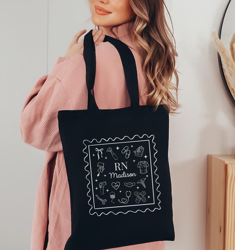 May include: Black tote bag with white line art of medical tools and the text "RN Madison" inside a decorative border. The bag has two black handles and is being held by a person wearing a pink top.