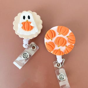May include: Two retractable badge reels with Halloween themes. One features a white ghost holding an orange pumpkin on a scalloped beige base. The other is a round white disc decorated with a repeating pattern of small, raised orange pumpkins.